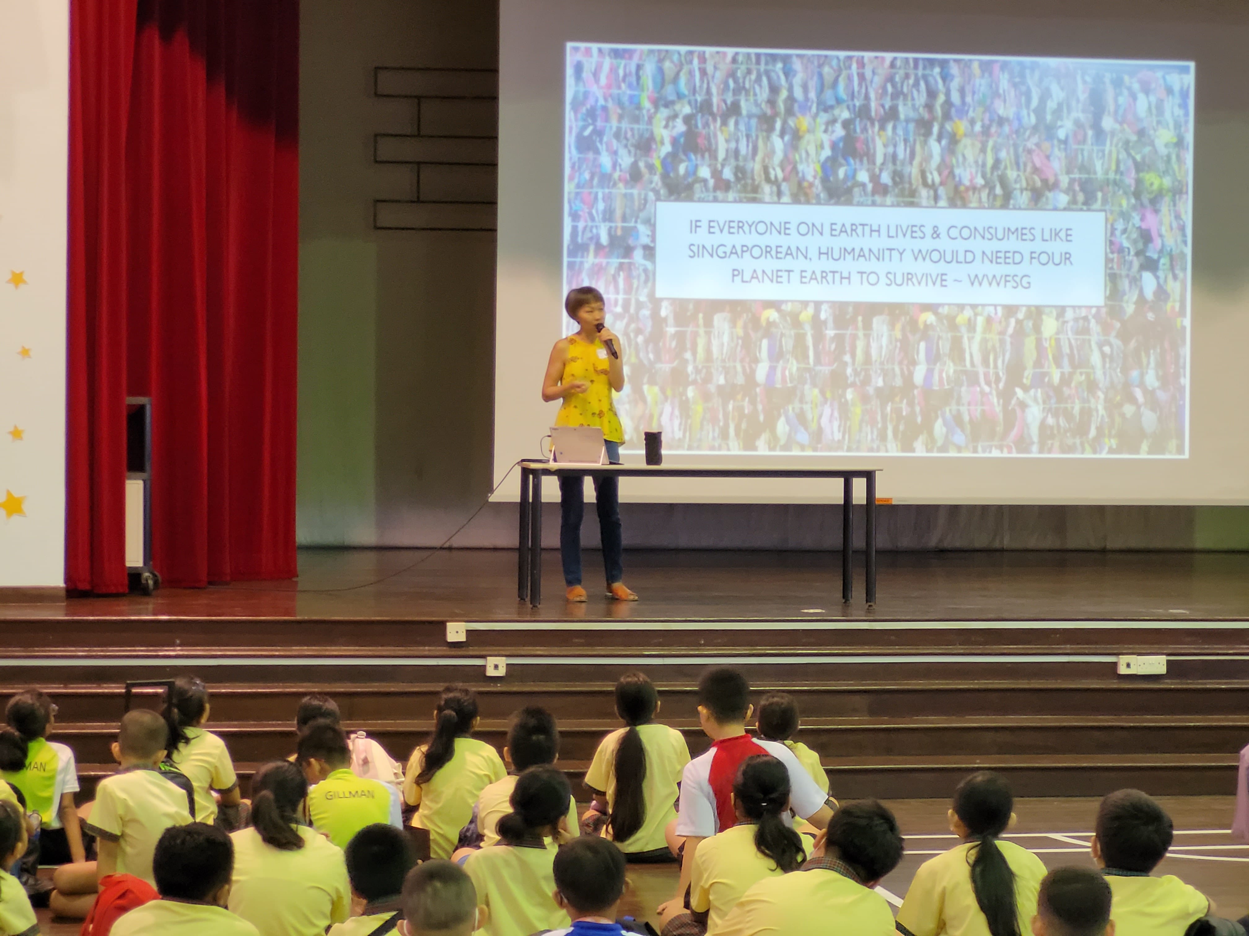 A woman in yellow speaks on stage to students in yellow shirts, with a sustainability-themed slide projected behind her.